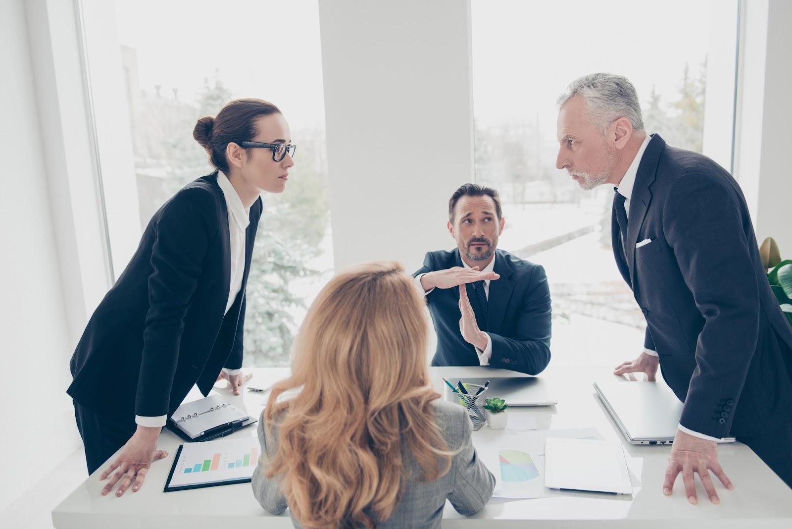 Parties to a contract dispute face each other aggressively across a conference table as a mediator holds up his hands in a “time-out” signal; concept image for resolving business conflicts. 