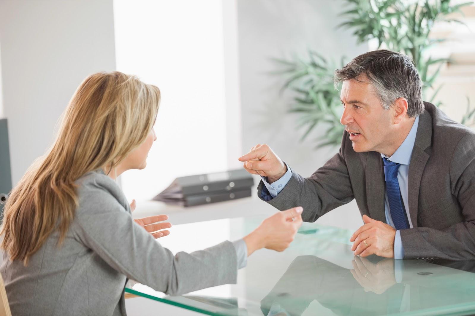 A blonde businesswoman and a dark-haired businessman arguing over a glass desk. 