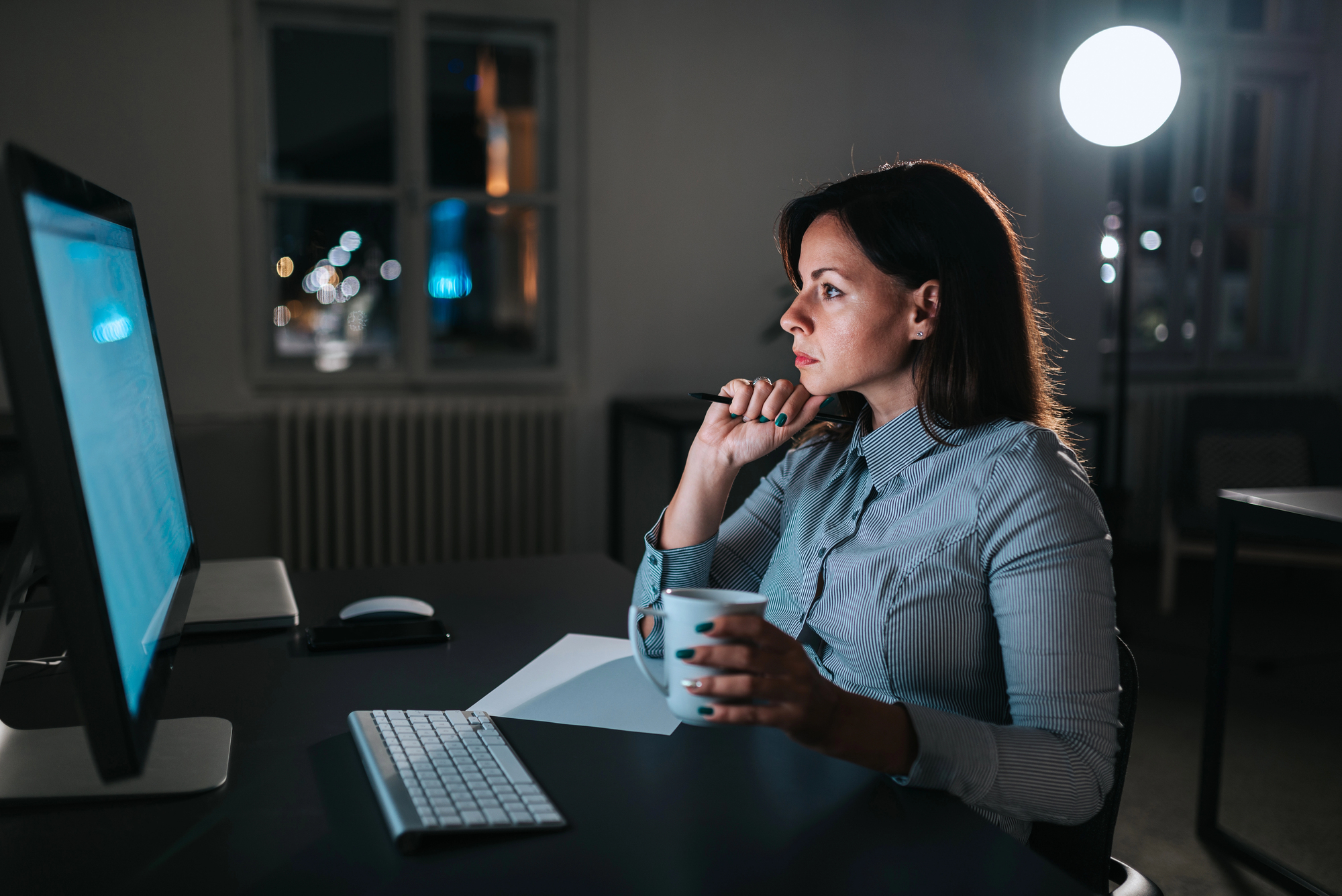  An employee drinking coffee in a darkened office, looking thoughtful as she reviews information on a computer screen. 