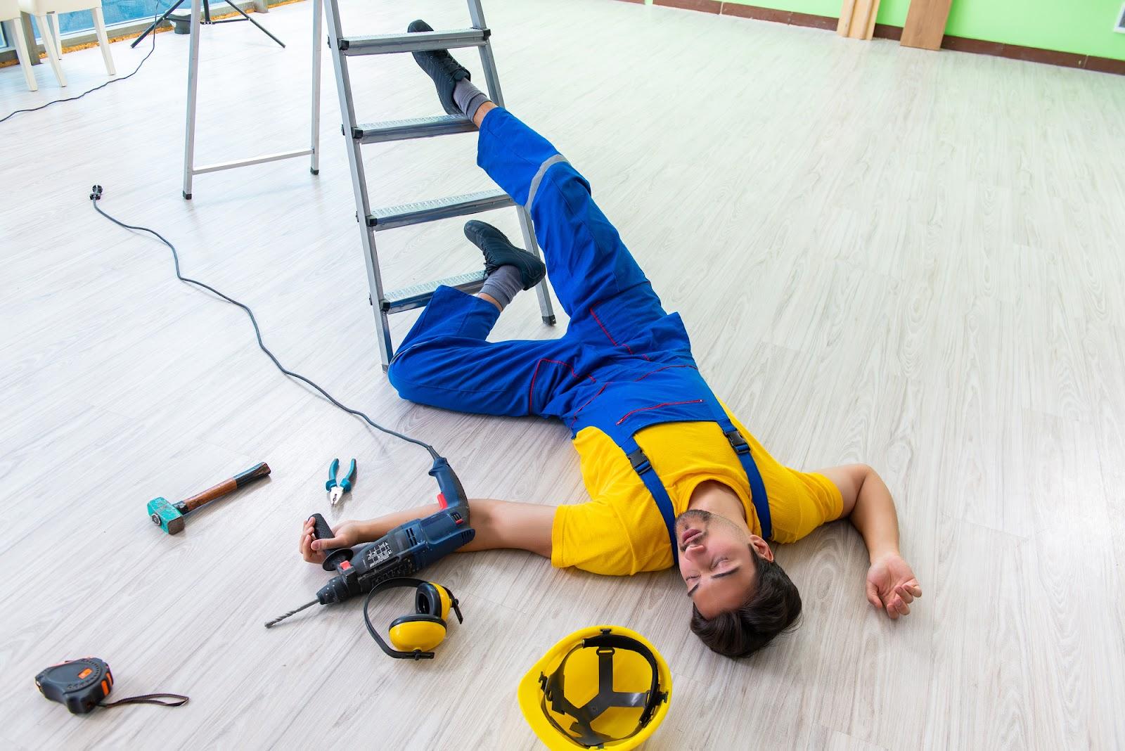 A dark-haired man dressed in blue overalls and a yellow shirt is lying on the floor, his legs tangled in a possibly defective ladder, a hard hat and assorted tools at his side. He may have a third-party liability claim in addition to Workers’ Compensation for this work-related injury.