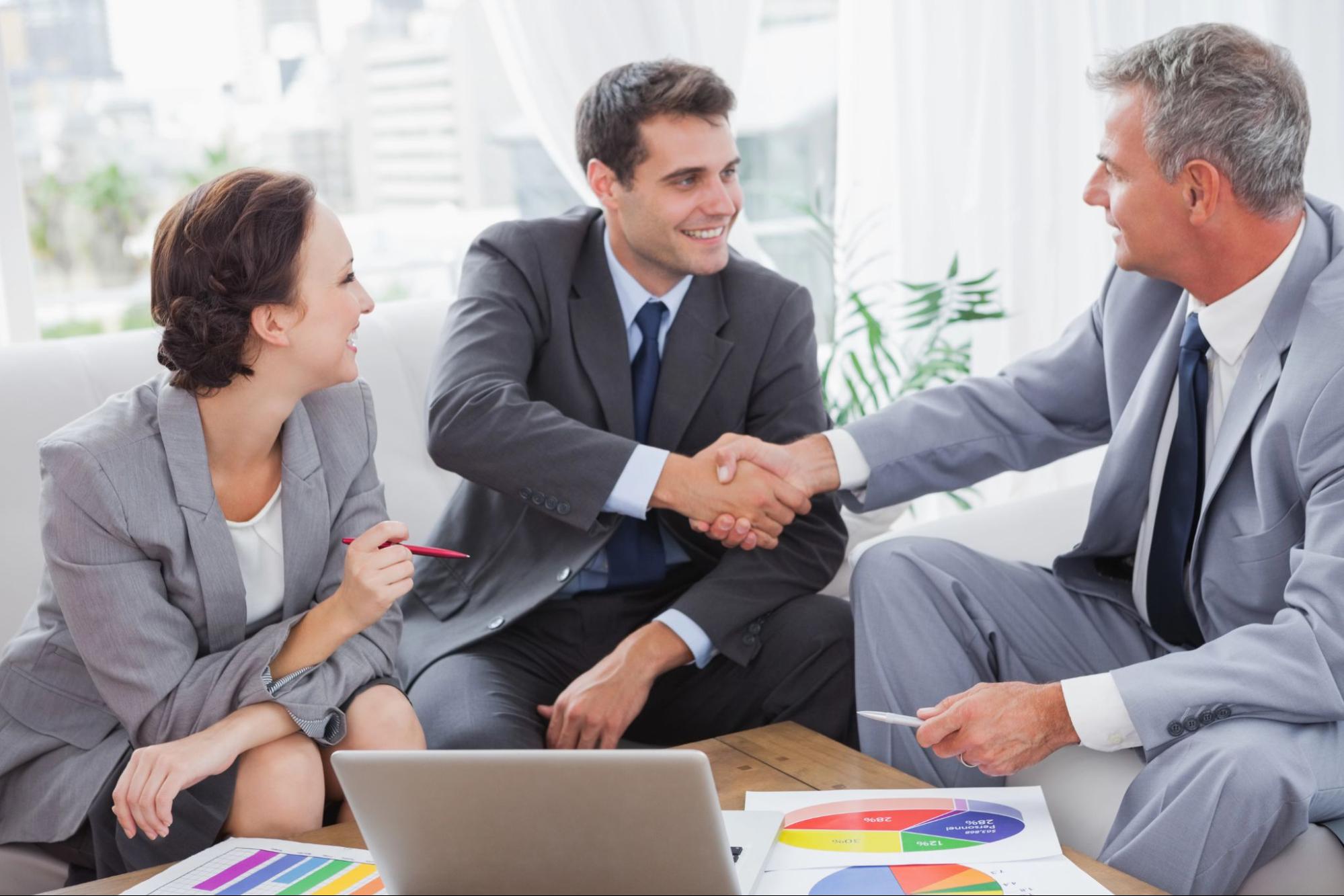 Three colleagues sitting around a low table with a laptop, reviewing pie charts and exchanging handshakes; forming relationships in business. 